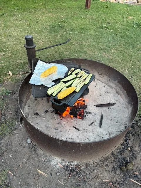 Corn cobs and zucchini cooking over an open campfire in a cast iron pan, flames glowing below.