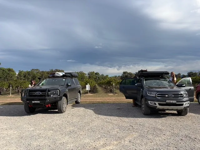Two 4WD vehicles with rooftop tents parked in a winery car park with vineyards and dramatic clouds behind.