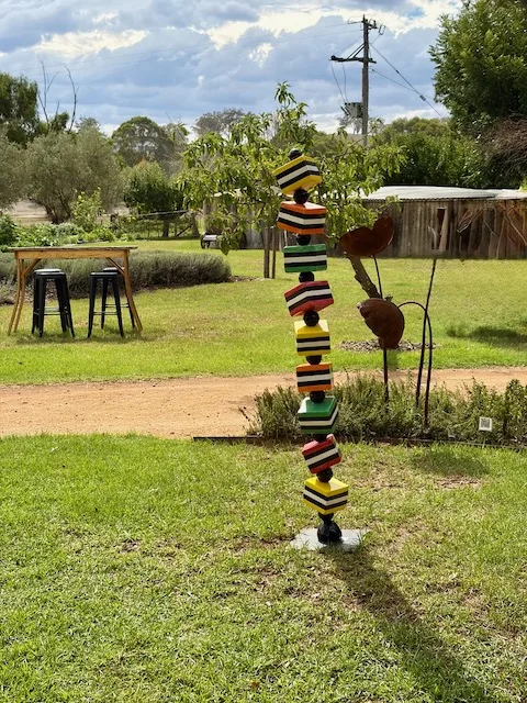 A colourful stacked geometric sculpture in the Rosby winery garden under a cloudy sky, with vines and buildings behind.