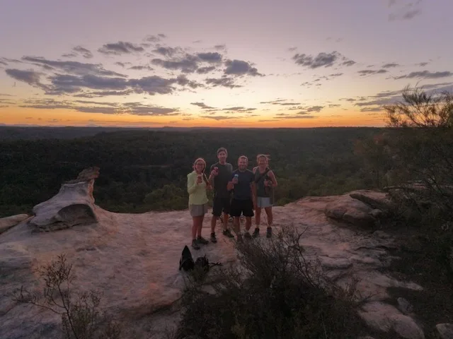 Four people standing on a sandstone ledge at the Pilliga Pottery viewpoint, drinks in hand, with a vast forested landscape stretching to the horizon under a deep purple and orange sunset sky.