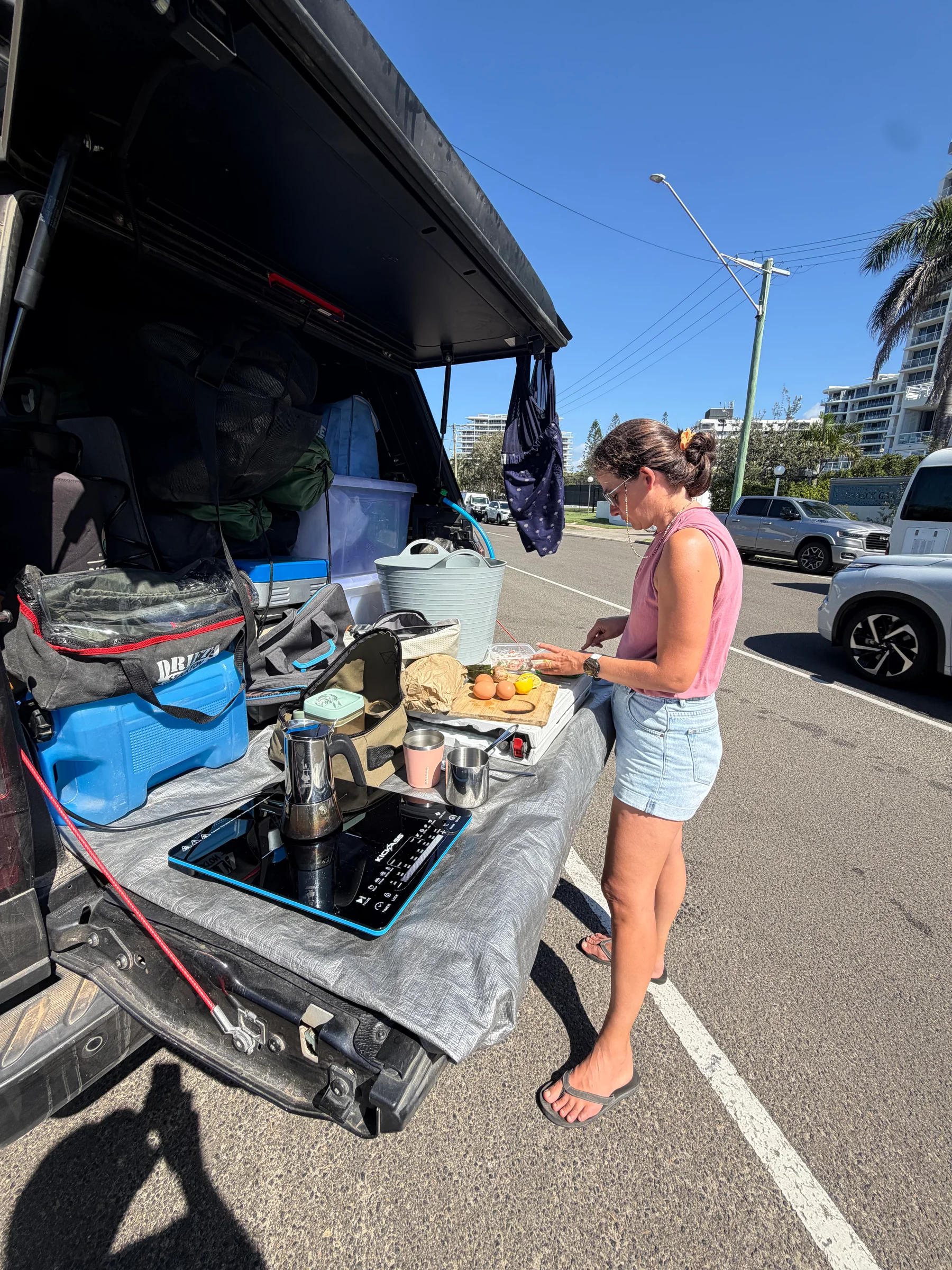 Steph preparing food at the open tailgate kitchen beside the Ranger on a sunny street.