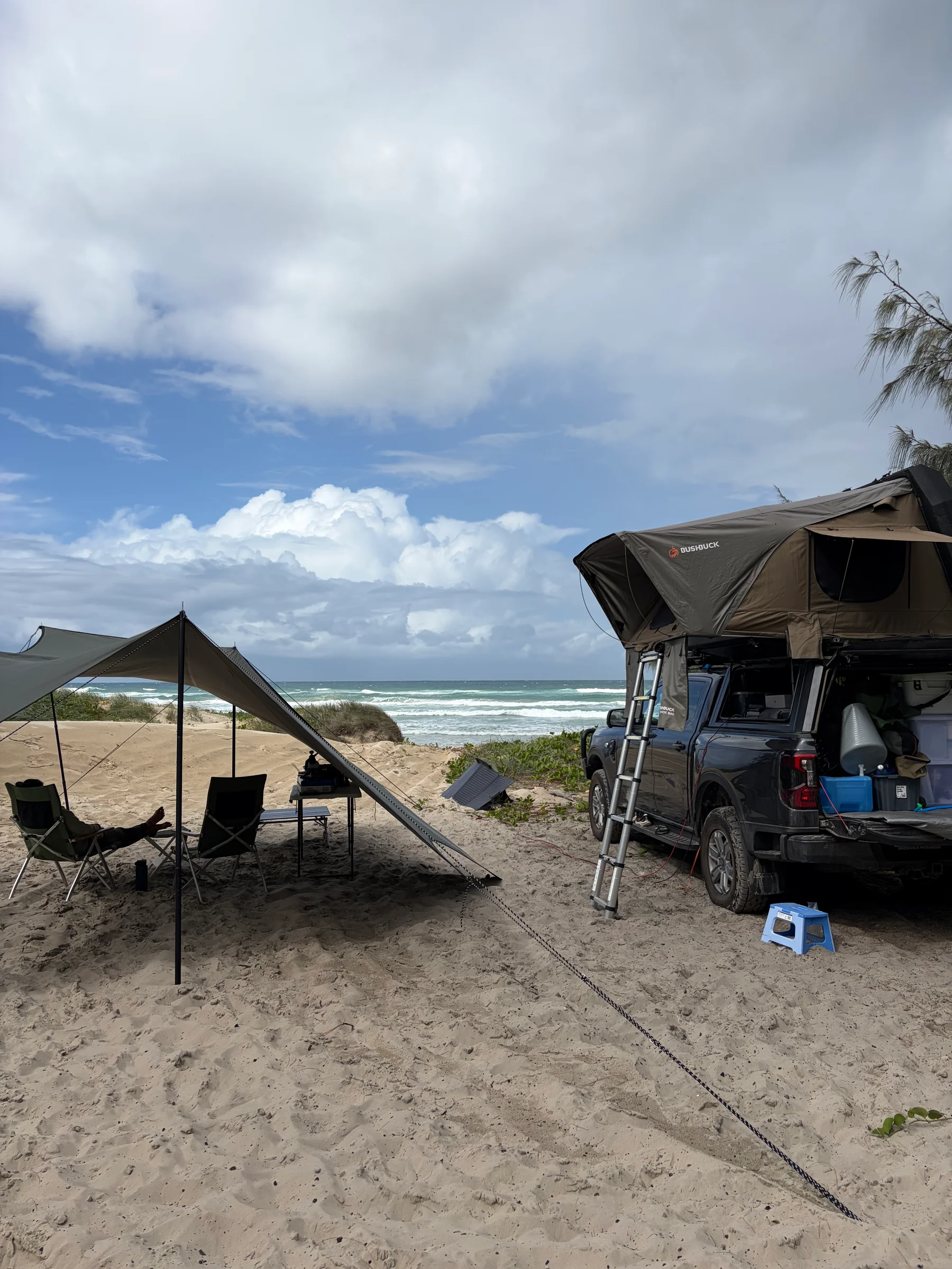 A white Land Cruiser with a rooftop tent parked on a wide beach, waves and overcast sky in the background.