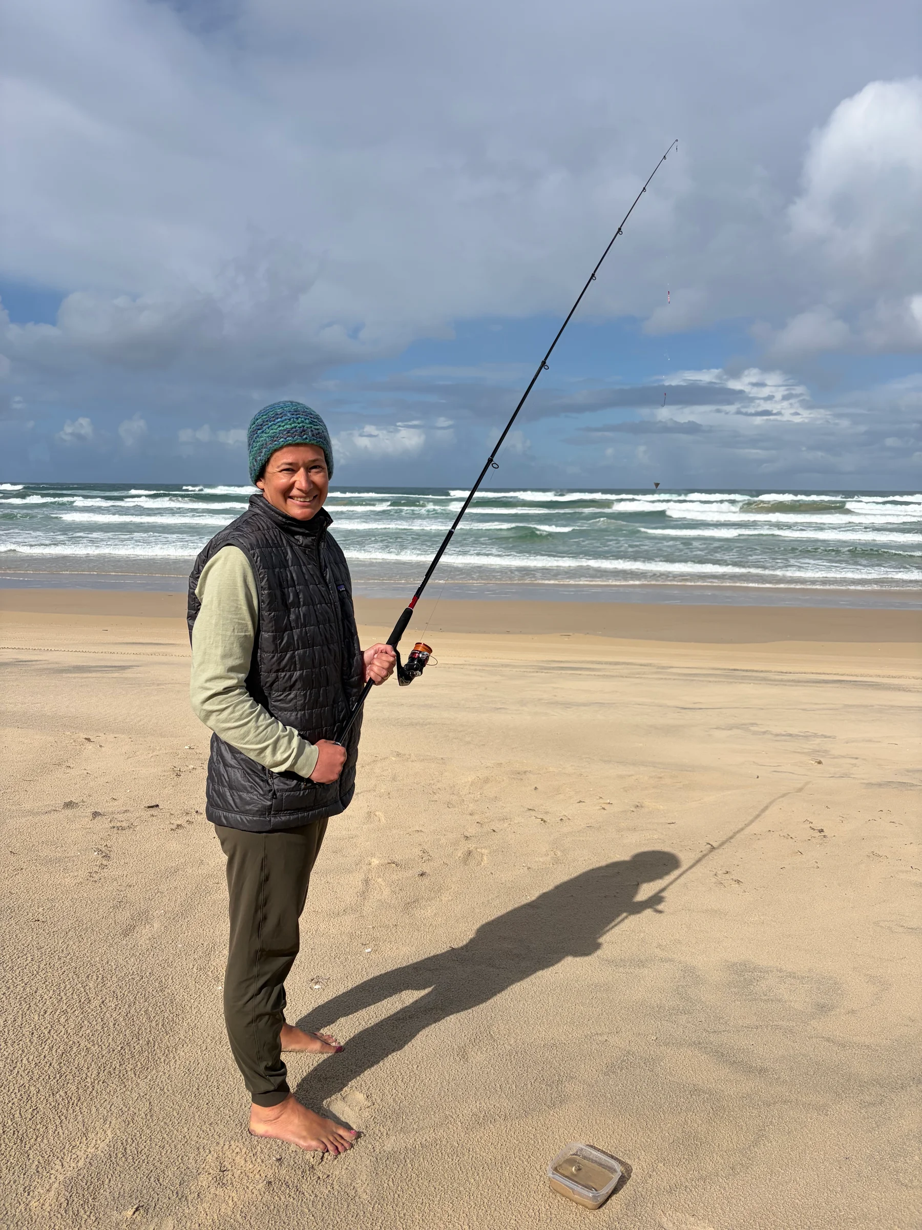 A person standing at the water's edge on Teewah Beach, holding a fishing rod, facing the ocean.