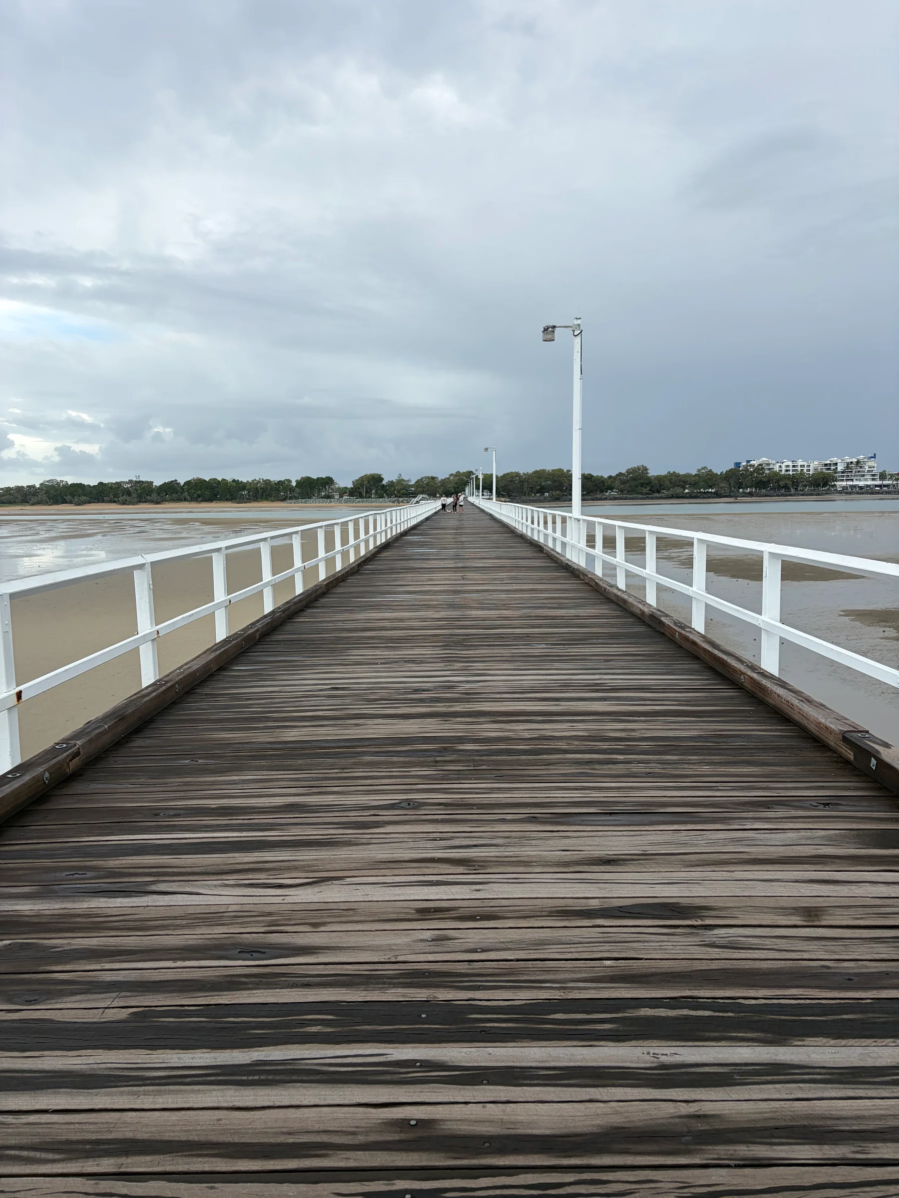 A long wooden jetty extending far out over calm blue water, the walkway narrowing into the distance under a clear pale sky.