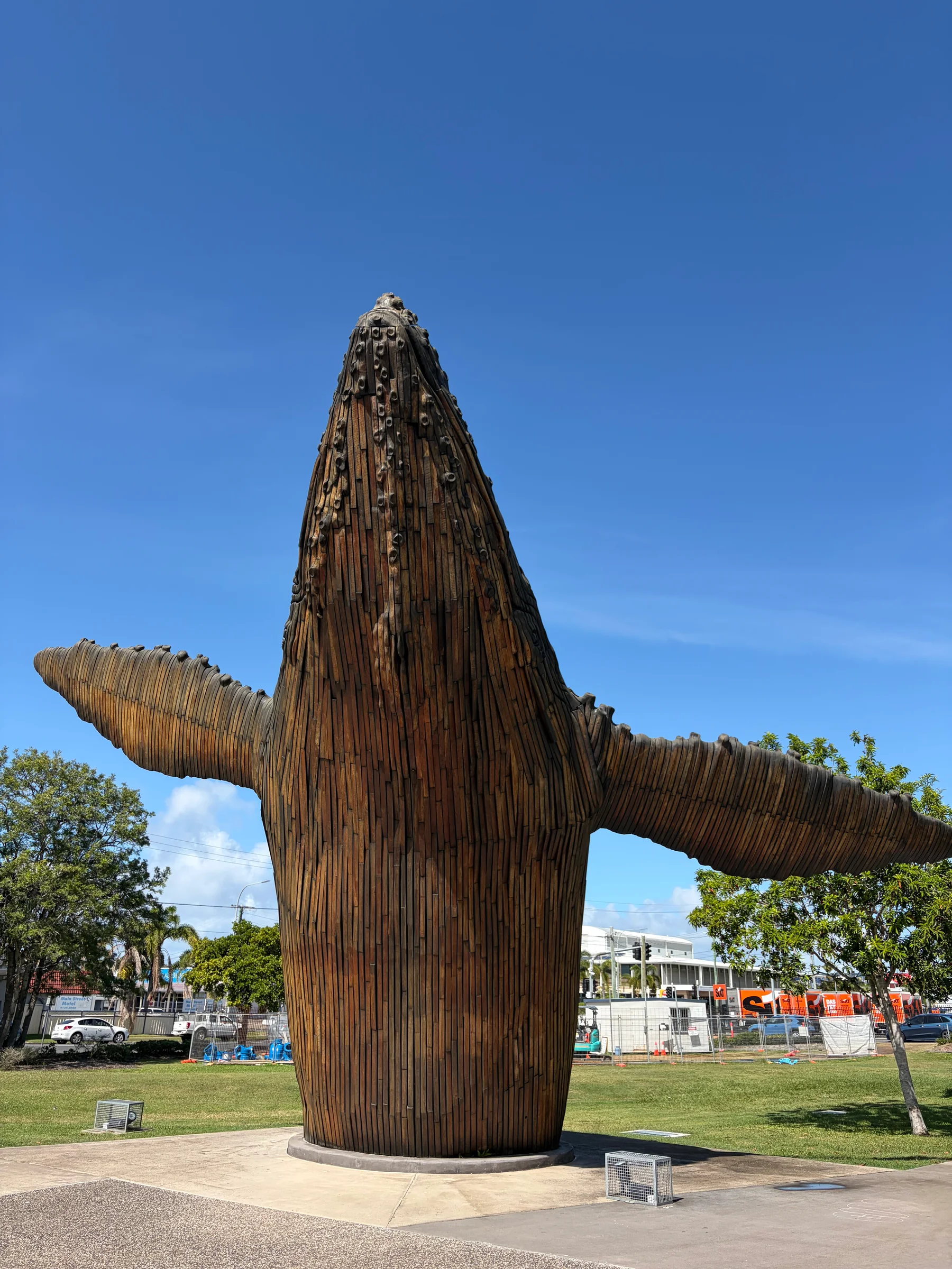 A large curved wooden sculpture resembling an animal form, standing in an outdoor public space surrounded by trees.