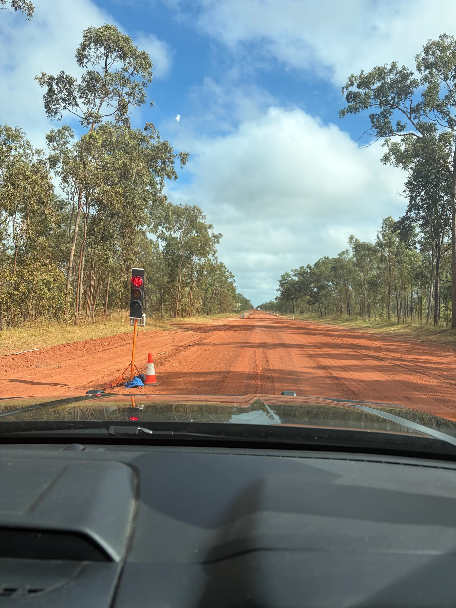 Red dirt road stretching into the distance with a temporary traffic light on red and a traffic cone, green bush on both sides