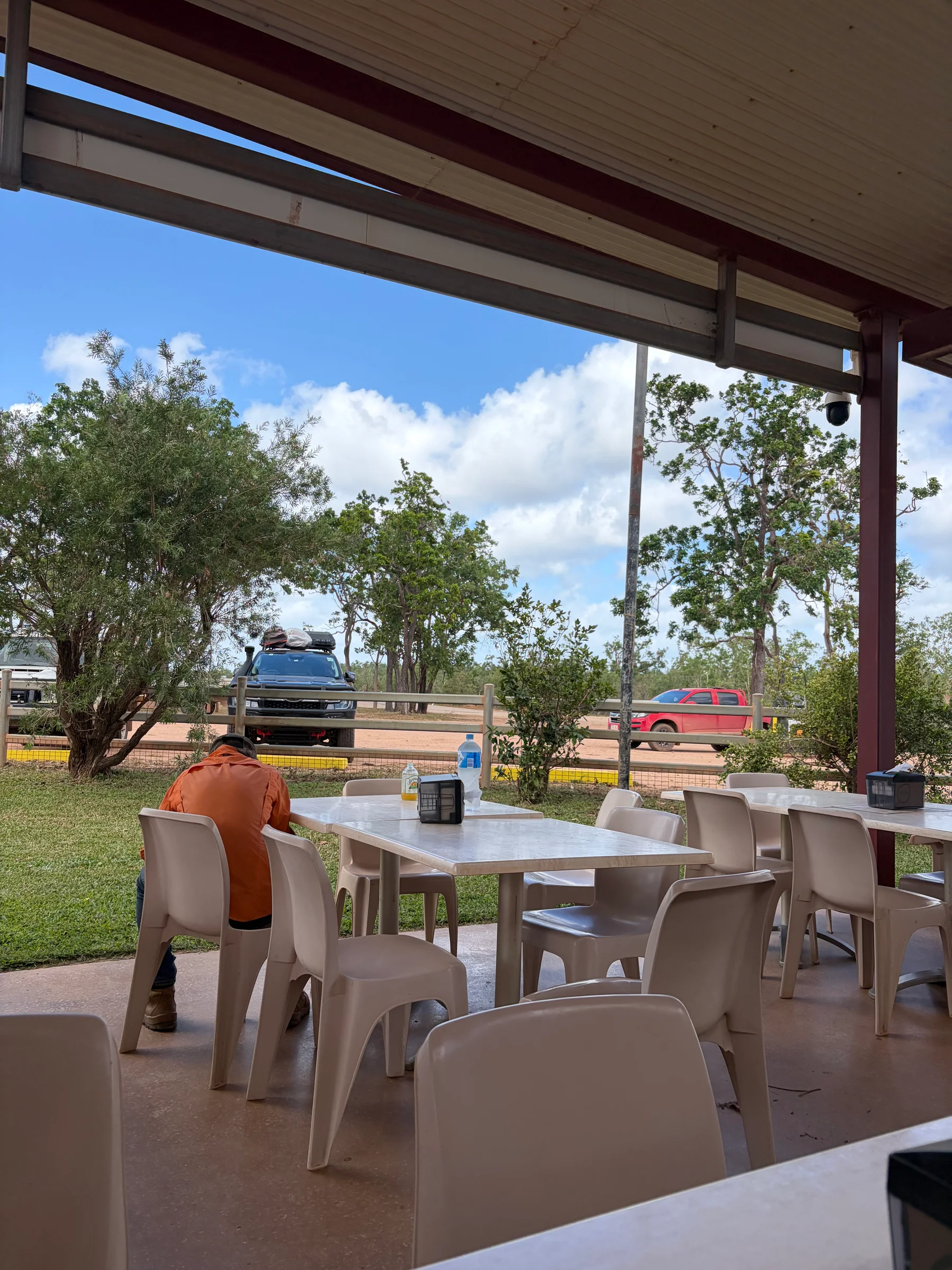 View from the shaded outdoor seating area at Archer River Roadhouse looking out to the carpark, Matt seated at a table in an orange shirt