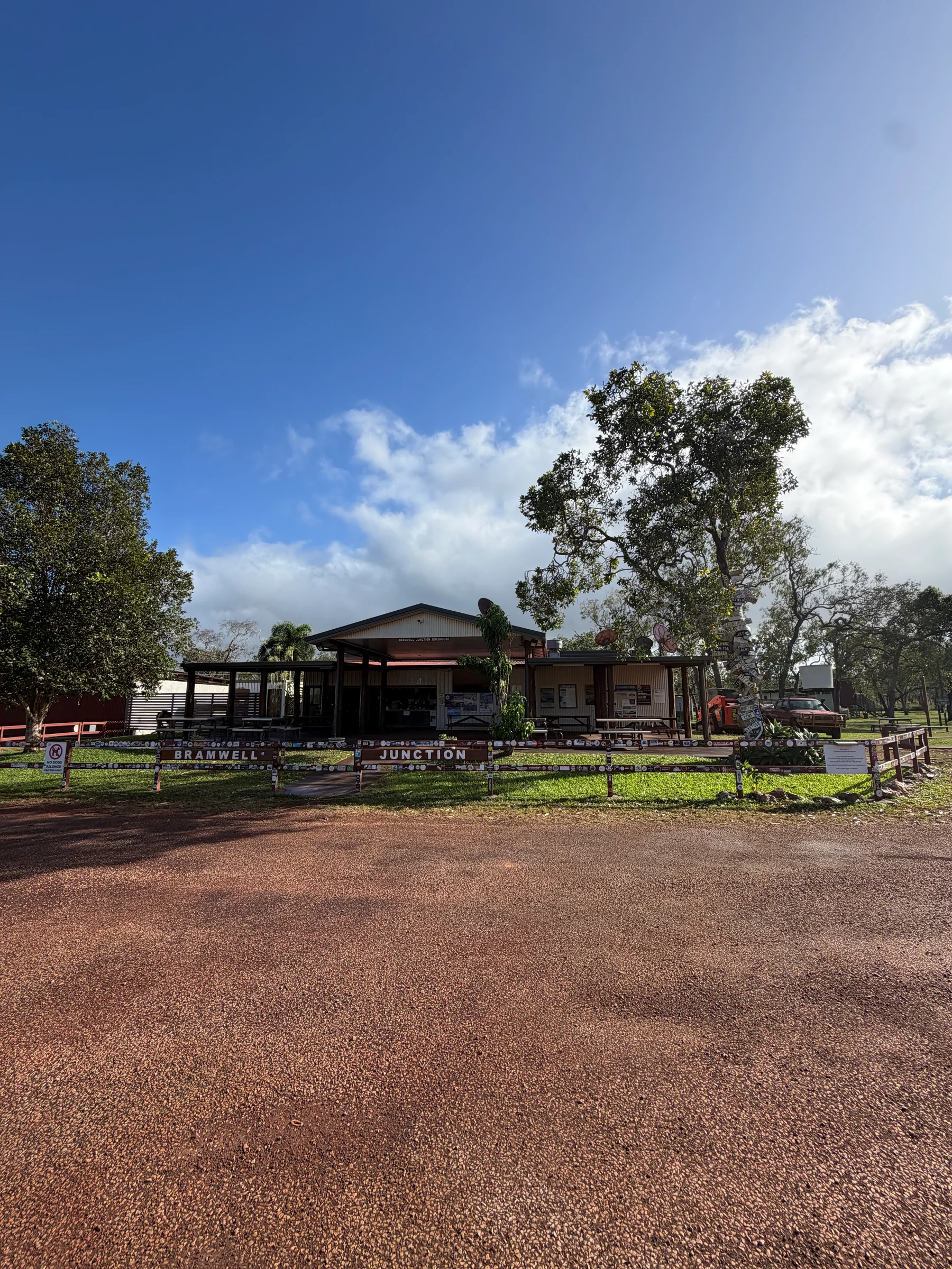 The Bramwell Junction Roadhouse building with the Bramwell Junction sign along a wooden fence, large gum trees above and red dirt in the foreground